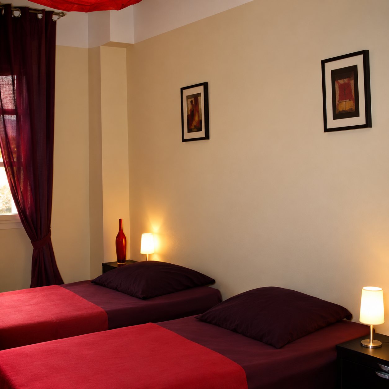 Une chambre aux murs beige clair avec deux lits recouverts de rouge, des rideaux violets, deux cadres au mur et des lampes allumées.