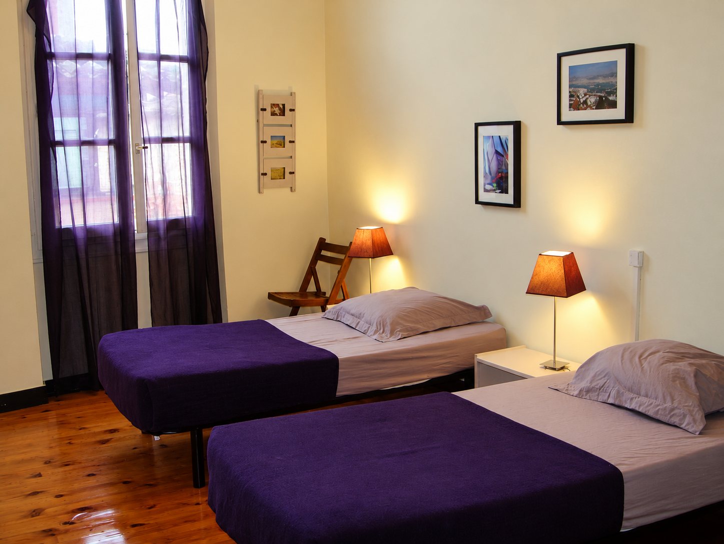 Chambre aux murs clairs avec deux lits simples couverts de violet, deux lampes allumées, des cadres au mur et une grande fenêtre aux rideaux violets.