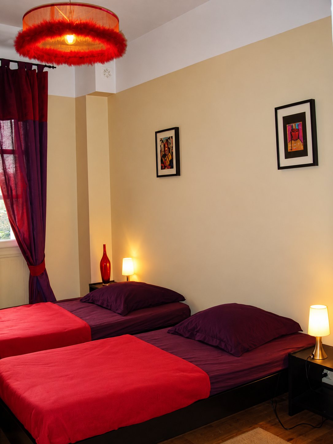 Chambre chaleureuse avec deux lits simples dressés de rouge et bordeaux, un lustre à franges rouges, des lampes de chevet allumées et deux cadres au mur beige.