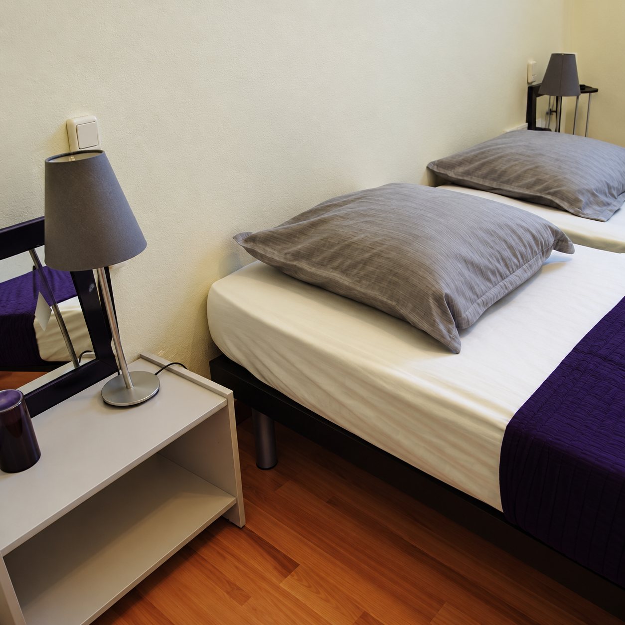 Une chambre aux murs clairs avec un lit habillé de gris et de violet, une table de chevet et une lampe grise.