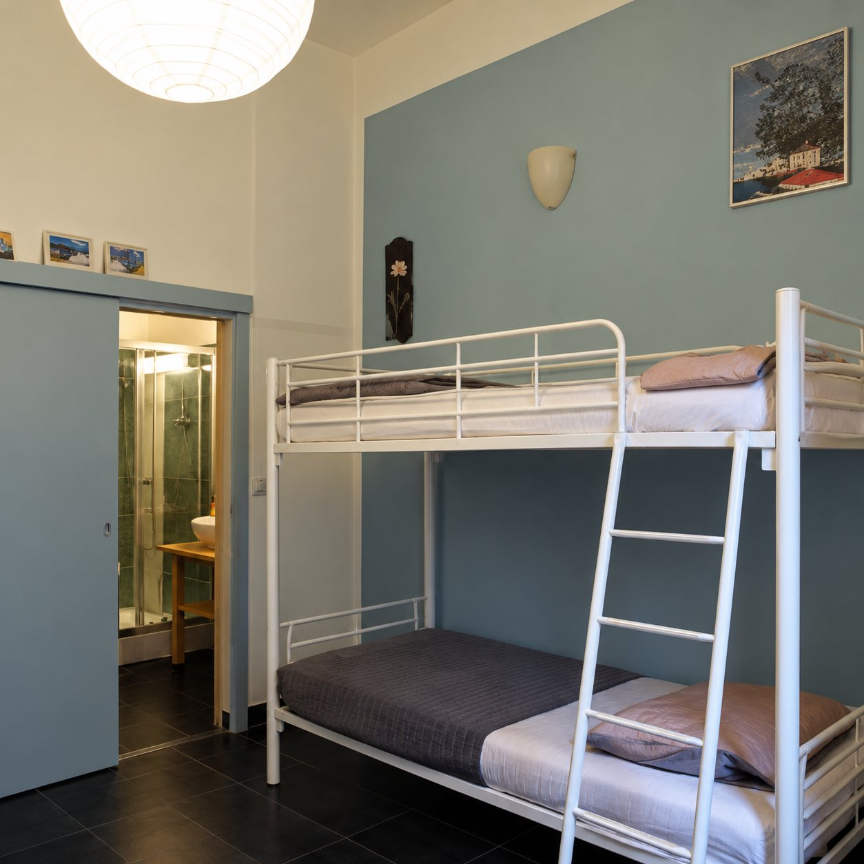 Une chambre simple avec des lits superposés métalliques blancs, un mur bleu clair, une suspension ronde et une porte ouverte vers une salle d'eau.