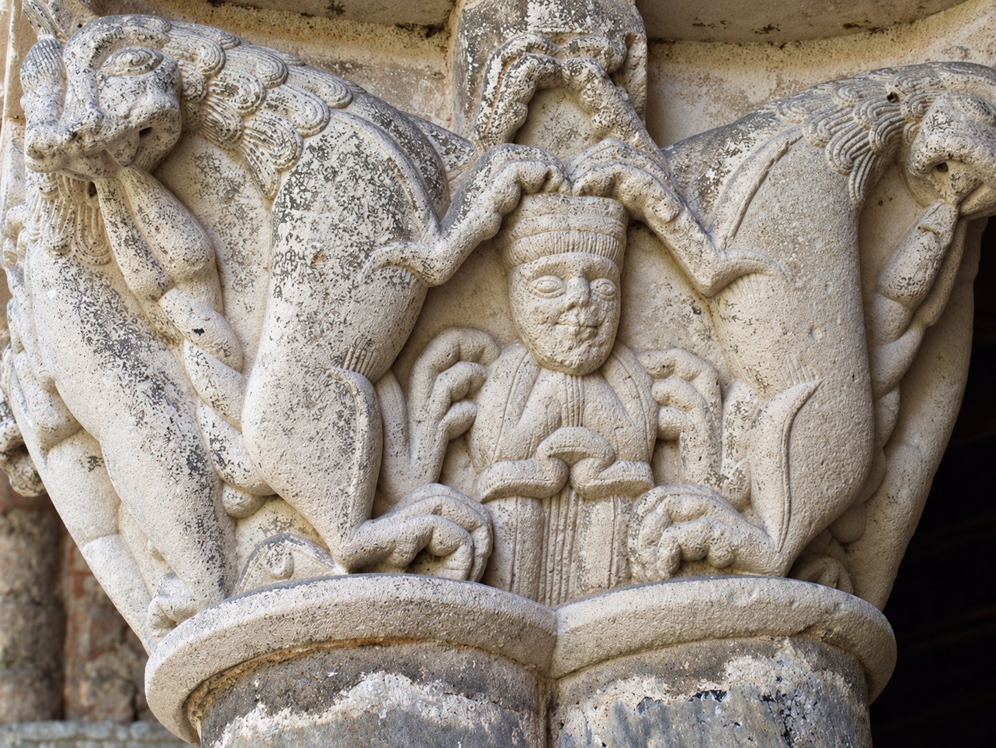 Gros plan sur un chapiteau de pierre romane sculpté représentant un personnage central encadré de deux animaux fantastiques dressés.