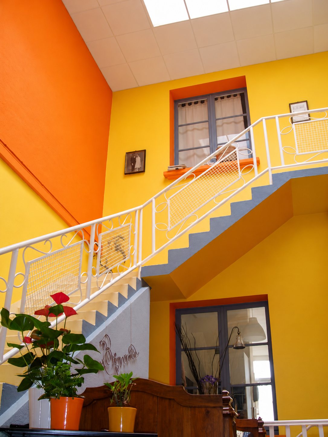 Un escalier à rampe blanche ouvragée monte dans un hall aux murs jaunes et orange, avec des plantes en pots au premier plan.