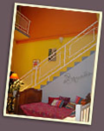 Un escalier blanc avec rambarde longe un mur jaune et orange au-dessus d'un lit aux coussins rouges.