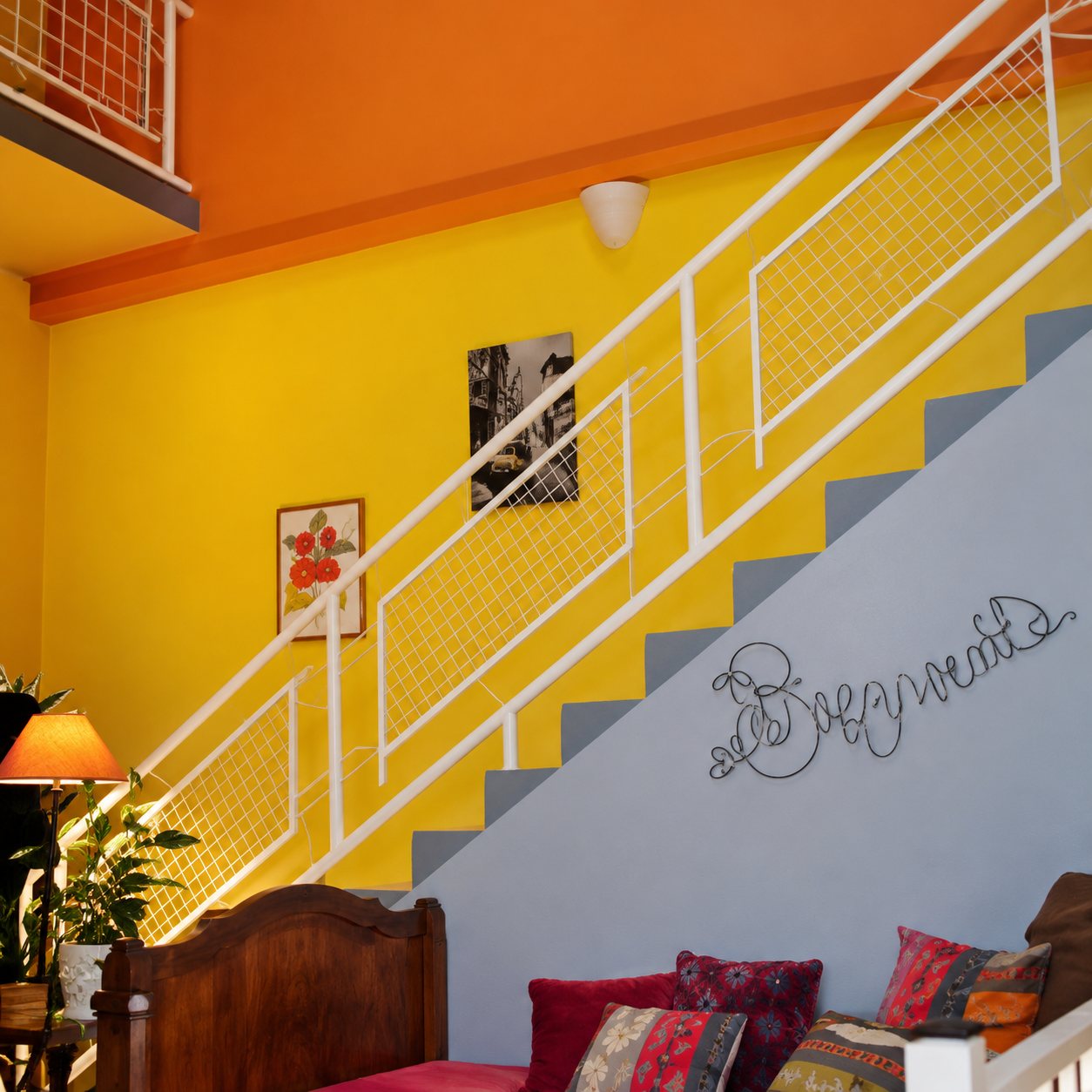 Un escalier à rampe blanche longe un mur jaune et gris dans un salon coloré, avec un canapé à coussins et une lampe au premier plan.