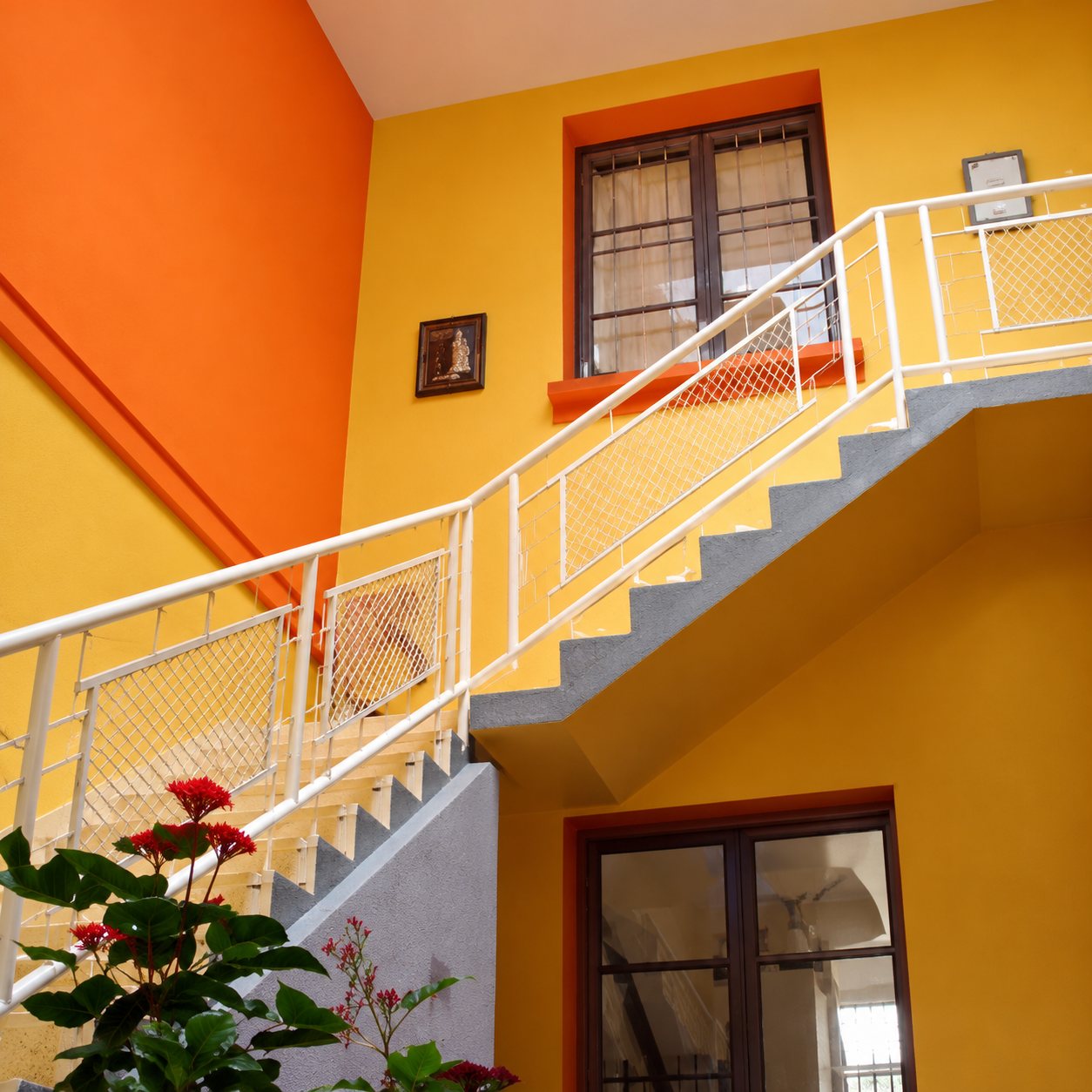 Un escalier intérieur à rampe blanche longe des murs peints en orange et jaune, avec une fenêtre en hauteur et une plante fleurie au premier plan.