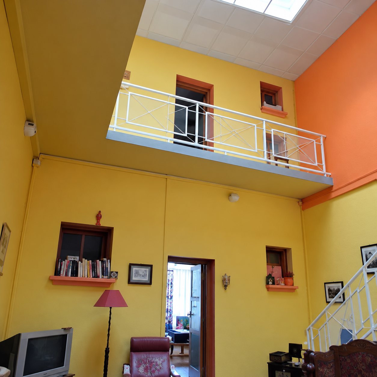 Pièce haute aux murs jaunes et orange, dotée d'un balcon blanc en mezzanine, d'une lampe et d'une porte ouverte sur une autre pièce.