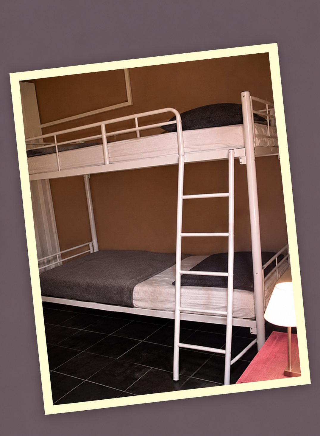 Un lit superposé en métal blanc avec échelle est installé dans une petite chambre aux murs sombres.