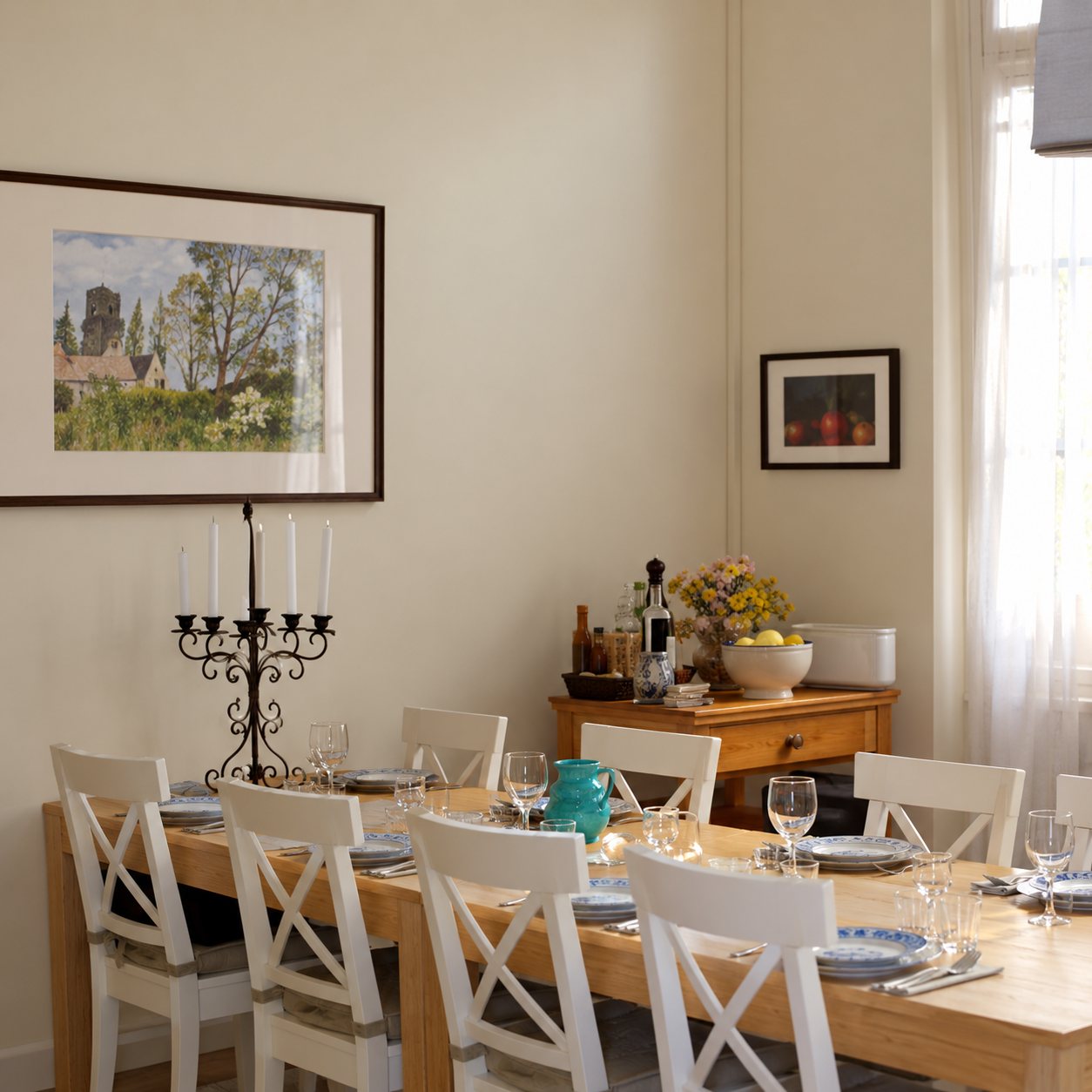 Une salle à manger claire avec une longue table dressée, des chaises blanches, un chandelier et des tableaux accrochés aux murs.
