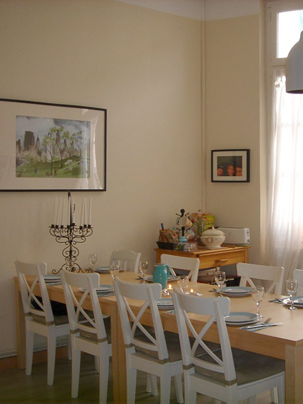 Une grande table en bois dressée avec assiettes et verres, entourée de chaises blanches à dossier croisé dans une salle à manger claire.