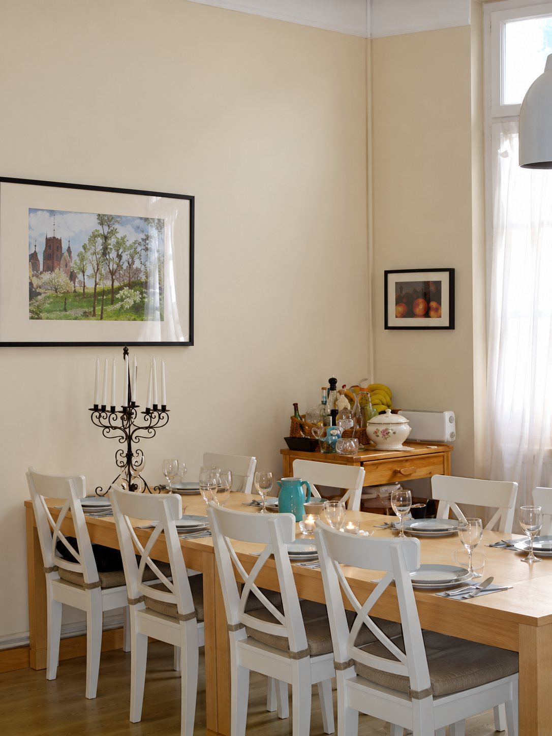 Une salle à manger claire avec une longue table dressée entourée de chaises blanches, un chandelier noir et des tableaux aux murs beiges.