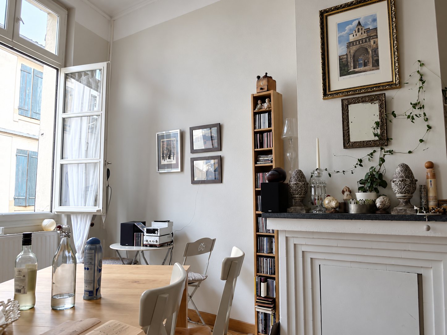 Photo en noir et blanc d'une pièce lumineuse avec une table et des chaises, une bibliothèque étroite, des cadres au mur et une cheminée décorée sous une fenêtre ouverte.