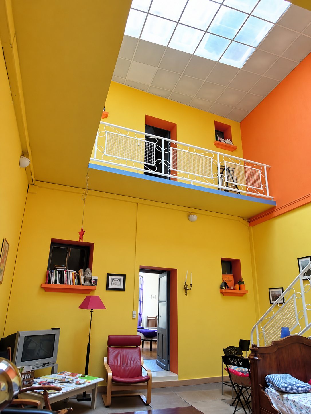 Salon aux murs jaunes et orange sous un puits de lumière, balcon blanc au premier étage